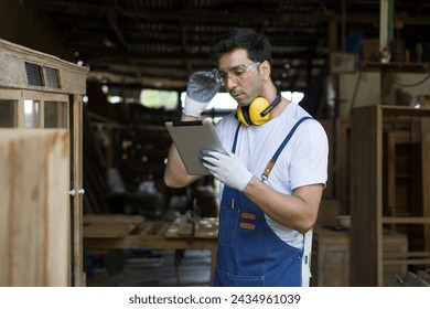 A carpenter or woodworker holding a tablet, reviewing design plan or instruction related to the woodwork. Wearing safety goggle, hearing protection headphone and glove stand in a woodworking workshop. - Powered by Shutterstock