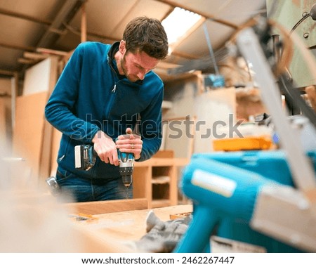 Carpenter At Workbench In Workshop Using Cordless Electric Drill