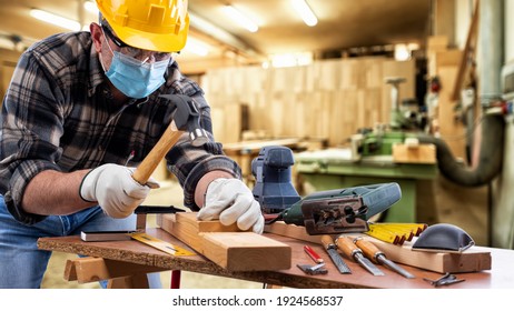 Carpenter worker at work in the carpentry workshop, wears helmet, goggles, leather gloves and surgical mask to prevent coronavirus infection. Preventing Pandemic Covid-19 at the workplace.