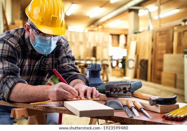 Carpenter worker at work in\
the carpentry workshop, wears helmet, goggles and surgical mask to\
prevent coronavirus infection. Preventing Pandemic Covid-19 at the\
workplace.