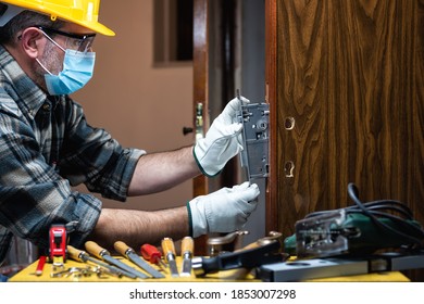 Carpenter Worker At Work Repair And Install A Room Door Lock, Wear The Surgical Mask To Prevent Coronavirus Infection. Preventing Pandemic Covid-19 At The Workplace. Carpentry.