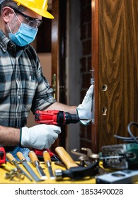 Carpenter Worker At Work Repair And Install A Room Door Lock, Wear The Surgical Mask To Prevent Coronavirus Infection. Preventing Pandemic Covid-19 At The Workplace. Carpentry.