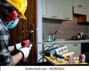 Carpenter Worker At Work Repair And Install A Room Door Lock, Wear The Surgical Mask To Prevent Coronavirus Infection. Preventing Pandemic Covid-19 At The Workplace. Carpentry.