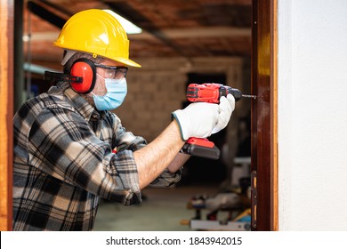 Carpenter Worker At Work Repairs And Installs A Room Door, Wear The Surgical Mask To Prevent Coronavirus Infection. Preventing Pandemic Covid-19 At The Workplace. Carpentry.