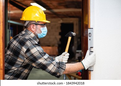 Carpenter Worker At Work Repairs And Installs A Room Door, Wear The Surgical Mask To Prevent Coronavirus Infection. Preventing Pandemic Covid-19 At The Workplace. Carpentry.