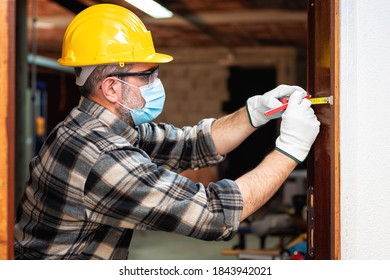 Carpenter Worker At Work Repairs And Installs A Room Door, Wear The Surgical Mask To Prevent Coronavirus Infection. Preventing Pandemic Covid-19 At The Workplace. Carpentry.