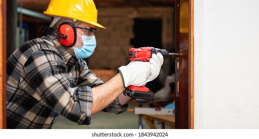 Carpenter Worker At Work Repairs And Installs A Room Door, Wear The Surgical Mask To Prevent Coronavirus Infection. Preventing Pandemic Covid-19 At The Workplace. Carpentry.