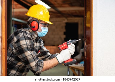 Carpenter Worker At Work Repairs And Installs A Room Door, Wear The Surgical Mask To Prevent Coronavirus Infection. Preventing Pandemic Covid-19 At The Workplace. Carpentry.