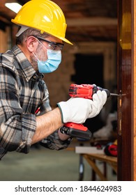 Carpenter Worker At Work Repairs And Installs A Room Door, Wear The Surgical Mask To Prevent Coronavirus Infection. Preventing Pandemic Covid-19 At The Workplace. Carpentry.