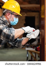 Carpenter Worker At Work Repairs And Installs A Room Door, Wear The Surgical Mask To Prevent Coronavirus Infection. Preventing Pandemic Covid-19 At The Workplace. Carpentry.