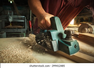 Carpenter working with electric planer on wooden plank in workshop.