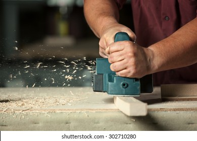 Carpenter working with electric planer on wooden plank in workshop.