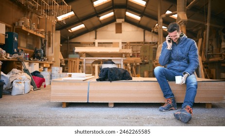 Carpenter Working In Woodwork Workshop With Pet Dog Making Call On Mobile Phone On Coffee Break - Powered by Shutterstock