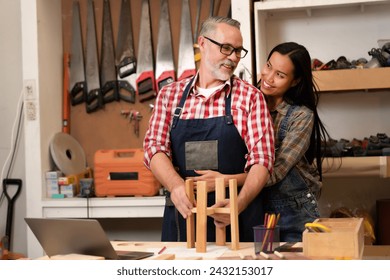 A carpenter's wife hugs her husband from behind to give him encouragement to work, feeling happiness with a smiling face at carpentry shop. Joiner, woodcraft, furniture, handmade, timber, wood