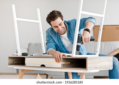 Carpentry, renovation work, furniture assembly, new purchase and hobby at home. Cheerful young busy handsome man screwing details to table with electric drill, in interior of living room, copy space