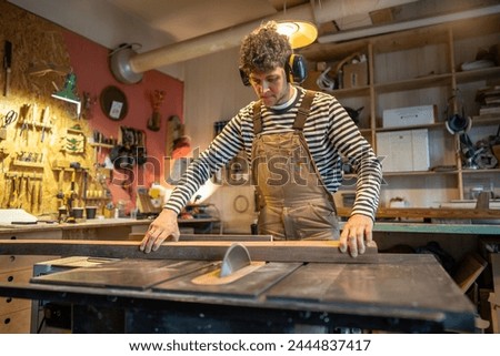 Carpentry workshop, focused craftsman master working . Thoughtful man artisan wearing earmuffs uses dangerous equipment, circular woodworking machine. Workplace safety, compliance. Woodwork, joinery.