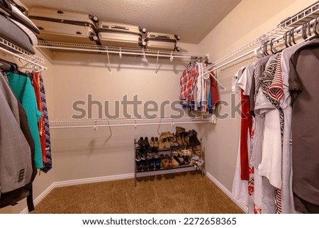 Carpeted walk-in closet with small to large suitcases above the bracket shelf. There is a wooden shoe rack on the right below the metal wire shelves with hanging clothes below.