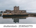 Carrickfergus Castle reflected in calm waters, with boats docked along the stone walls. The medieval fortress stands against a cloudy sky in Northern Ireland harbor