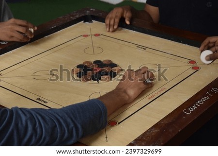 a carrom game in progress. Players’ hands flick the striker on a traditional Indian board. The wooden board with white and black pieces is the focus The game embodies culture, leisure, and strategy.