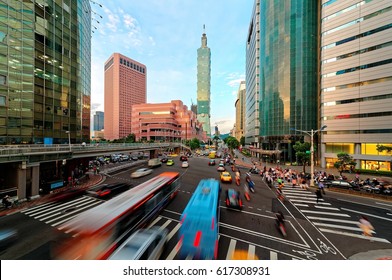 Cars, buses and scooters dashing by a busy street corner and people passing on the crosswalk in Downtown Taipei at rush hour, with World Trade Center building and 101 Tower in Xinyi Financial District