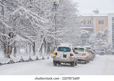 Cars Drive Along A Snow-covered City Street. Heavy Snowfall And Snowstorm In The City. A Lot Of Snow On The Roadway, Cars And Trees. Cold Snowy Winter Weather. Magadan, Siberia, Russia.