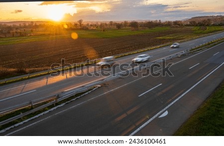 Cars drive at high speed on the highway through the rural landscape. Fast blurred highway driving. A scene of speeding on the highway. Beautiful sunset in the background.