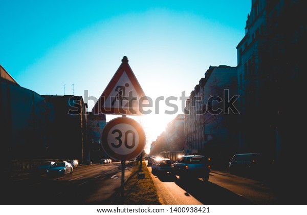 Cars driving down a city street\
at sunset with the bright flare of the sun behind a road works\
speed sign in the center of the road and reflection on the\
tarmac