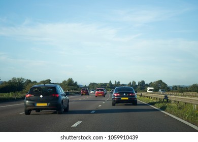 Cars Driving Down Motorway In Summer Time Alongside Countryside.