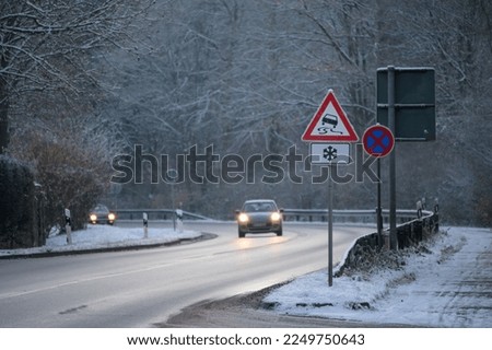 Cars driving down a wet mountain road in winter with a slippery road and ice warning sign covered in snow and ice and snow on the sides of the road and on trees
