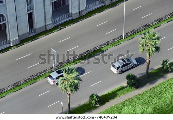 Cars go by road in the city\
street divided by green spaces and palm trees. View from\
above.