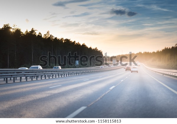 Cars in motion blur on highway during sunset. big\
road with metal safety barrier or rail under the cloudy blue sky\
and yellow and orange sun.