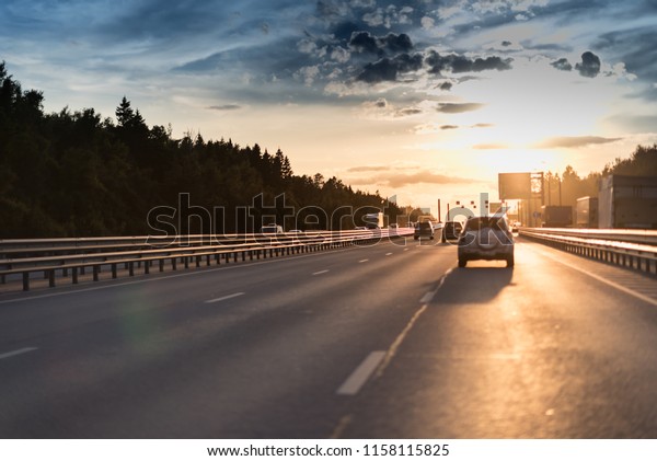 Cars in motion blur on highway during sunset. big\
road with metal safety barrier or rail under the cloudy blue sky\
and yellow and orange sun.