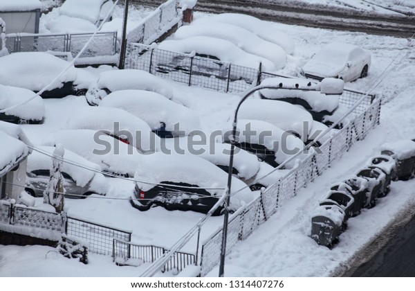 Cars on parking and street covered with big snow\
layer. View of winter and snowing on city street with snowflakes.\
In snowy season, motor vehicles with lot snow because of snow\
drifting in winter day.