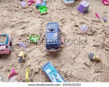 Cars and other plastic toys are scattered on the beach sand.  Soft focus.