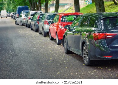 Cars Parked Along Narrow City Street. Row Of Parked Cars. Many Cars Parked And Lined Up Under Trees On Urban Street. Public Parking Lot