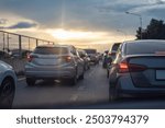 Cars stuck in traffic on a city highway during sunset, with brake lights glowing under a cloudy sky, creating a moody atmosphere.