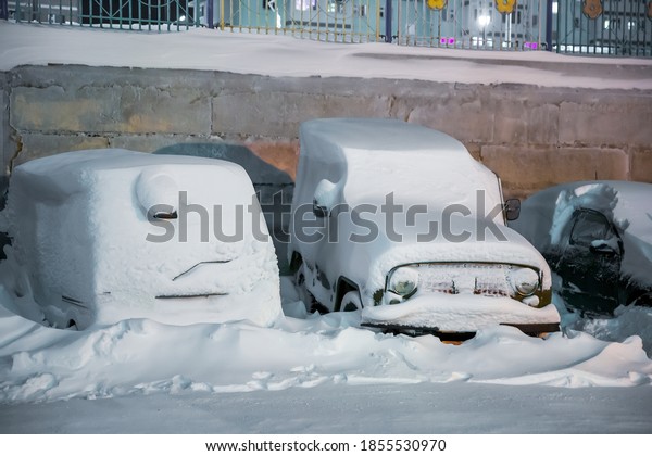 Cars under the snow after a snowfall. There is a\
lot of snow on the roofs, windows and hoods of cars. Large\
snowdrifts on the street. Cars in a snow-covered parking lot. Cold\
snowy winter weather.