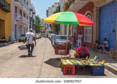 Cartagena, Columbia -- April 21, 2018. An Artist Works On A Painting Under An Umbrella; A Delivery Man Walks On A Side Street In Cartagena, Columbia.  Editorial Use Only.