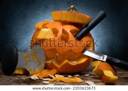 Carved Halloween pumpkin, Jack Lantern with carving tools. Spooky laughing, scary head. Jack-o'-lantern face cut out with spoon gutter, saw blade carvers. Seeds and pieces scattered around.