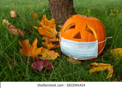 Carved Halloween Pumpkin With Sad Eyes Wearing Mask On The Grass With Leaves, No Party During Covid Or Coronavirus Outbreak, Events And Parties Canceled To Prevent The Risk Of Spreading The Virus