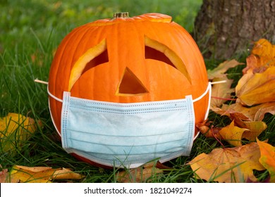 Carved Halloween Pumpkin With Sad Eyes Wearing Mask On The Grass With Leaves, No Party During Covid Or Coronavirus Outbreak, Events And Parties Canceled To Prevent The Risk Of Spreading The Virus