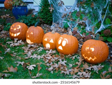 Carved pumpkins with different faces, displayed on the ground surrounded by leaves and greenery, creating a festive Halloween decoration scene.