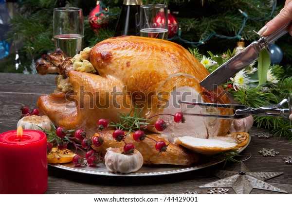 Carving rustic style roasted Christmas turkey
garnished with roasted garlic, lemon, and rosehips. Surrounded with
rustic Christmas ornaments, candles, wine, flowers, and Christmas
tree.
