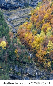 A cascading waterfall flows down a rugged rocky slope, framed by vibrant autumn foliage in a tranquil valley setting.