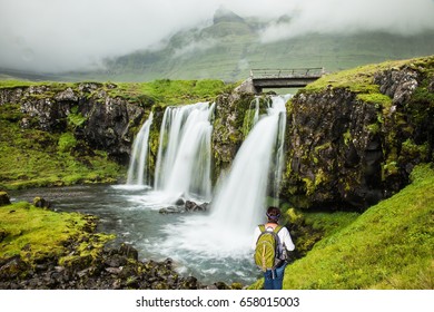 Cascading Waterfall Kirkjoufellfoss. Summer In Iceland. Elderly Woman -  Traveler With A Large Backpack Admiring The Waterfall. Concept Of Exotic And Extreme Tourism 