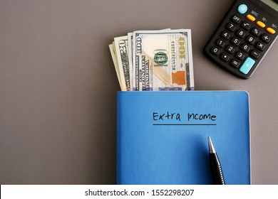 Cash dollar money ,a pen, calculator on a blue notebook with text written on cover EXTRA INCOME, on grey background, concept of making more money with writing or freelance job