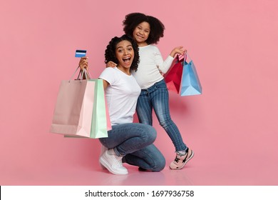 Cashback Concept. Portrait of cheerful african mom and her cute daughter holding credit card and colorful shopping bags, looking at camera