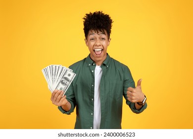Cashback, loan, giveaway. Rich wealthy excited handsome young black guy in casual outfit holding money cash dollar banknotes in his hand, showing thumb up over yellow studio background, copy space