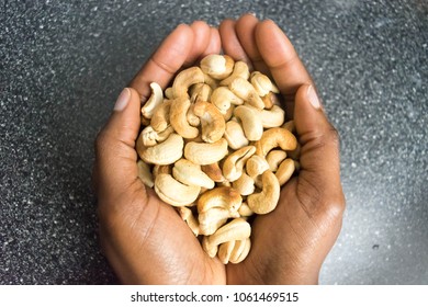 Cashew Nuts In The Cupped African American Hands Isolated On Dark Counter Top For Healthy Oils And Nuts And Dieting Concept. Cashew, Hazelnuts, Peanuts And Walnuts In The Palms.