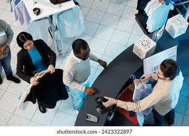 Cashier at clothing store checkout desk scanning and processing purchase for customer top view. Diverse people standing in line at counter desk while boutique employee using scanner - Powered by Shutterstock